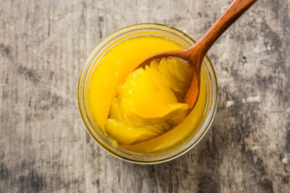 Wooden spoon scooping ghee from glass jar on rustic table Clarified butter or ghee in open glass jar with wooden spoon on rustic wooden background
