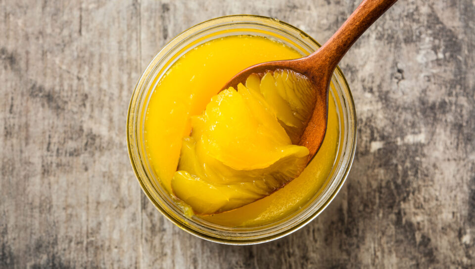 Clarified butter or ghee in open glass jar with wooden spoon on rustic wooden background