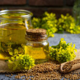 Mustard oil in a glass jar and decanter, mustard grains on a bur Mustard oil in a glass jar and decanter, mustard grains on a burlap napkin, flowers and leaves on wooden board background